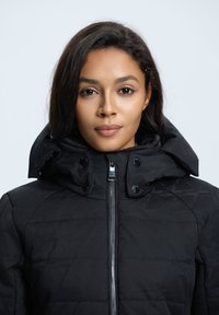 Schwarze gesteppte Jacke mit hohem Kragen, auffälliger Kapuze, Frontreißverschluss und Druckknopfverschluss. Subtile geometrische Musterstruktur.