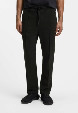 Mężczyzna ubrany w czarne spodnie o prostych nogawkach ze ściągaczem w talii i czarne trampki, stojący na jednolitym białym tle.