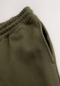 Alyvuogių žalios spalvos sportinės kelnės su elastine juosmens juosta, šonine kišene ir minkštu audinio paviršiumi. Dizainas yra paprastas ir kasdieniškas.