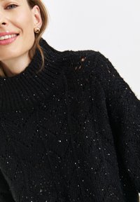Czarny dziany sweter z wysokim kołnierzem, z fakturowanym wzorem i subtelnymi cekinami, które tworzą efekt połysku.