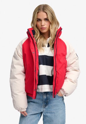 Blond kvinde iført en rød og hvid dunjakke, stribet skjorte og blå jeans, står med den ene hånd i lommen mod en hvid baggrund.