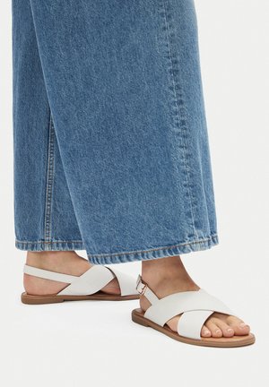 Białe płaskie sandały z krzyżującymi się paskami, regulowaną klamrą i płaską podeszwą, w połączeniu z jasnoniebieskimi szerokimi dżinsowymi spodniami.