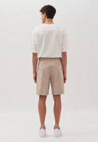 Pantaloni corti beige con tasche posteriori, orlo arrotolato, abbinati a una t-shirt bianca morbida. Modello che indossa sneakers bianche. Sfondo neutro.