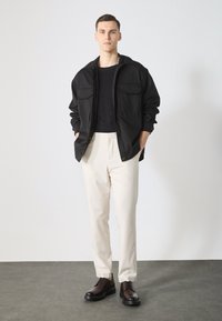 Men's outfit featuring a black jacket with two chest pockets, a black shirt, cream trousers, and brown lace-up shoes on a neutral background.
