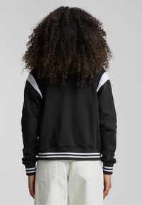 Črna majica s kapuco z belimi poudarki na ramah, rebrastimi manšetami in pasom. Mehka tkanina, sproščen kroj. Kombinirana s svetlimi hlačami.