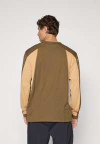 En mann som ser bortover, iført en to-tone langermet skjorte i olivengrønn og beige med tekst på høyre erme, sammen med svarte bukser.