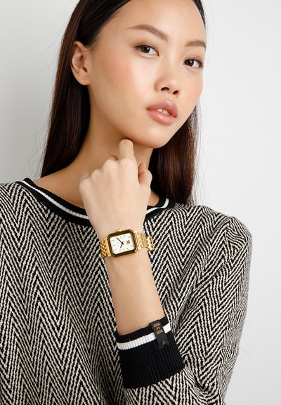 Giovane donna che indossa un maglione a motivo bianco e nero, mostrando un orologio da polso dorato con quadrante quadrato sul polso sinistro sollevato.
