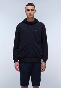 Felpa con zip di colore blu scuro, dotata di tasca a marsupio, cappuccio con cordini e piccolo logo sul petto. Abbinata a pantaloni corti blu navy con tasche cargo.