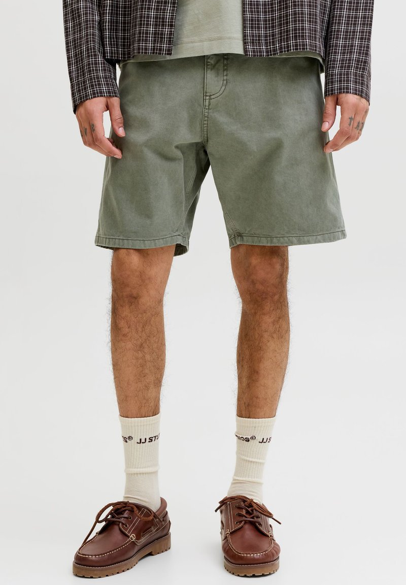 Άνδρας που φοράει χακί σορτς, εκρού κάλτσες με κείμενο, καφέ δερμάτινα παπούτσια με κορδόνια και ένα καρό σακάκι πάνω από πράσινο πουκάμισο.