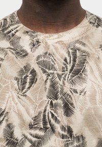 Gros plan d'une personne à la peau foncée portant une chemise beige avec des motifs de feuilles abstraites noirs et bruns et un col rond.