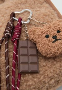 Breloczek z teksturowanym charms z brązowym misiem obok zawieszki w kształcie czekolady, przymocowany czerwonymi i brązowymi plecionymi sznurkami oraz metalowym zapięciem.