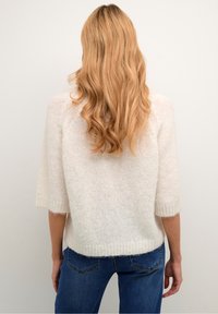Maglione bianco, strutturato, con maniche corte e una vestibilità ampia, abbinato a jeans blu. Il maglione ha un aspetto morbido e soffice.