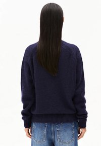 Femme aux longs cheveux noirs et lisses portant un pull marin en maille bleu marine et un jean bleu, vue de dos sur un fond blanc.