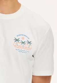 Baltas īsajām piedurknēm T-krekls ar grafiku, kurā attēloti palmu koki, kalni un saule oranžā un zaļās krāsas tonī, ar uzrakstu "Antigua Port Beach Club".