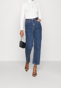 Blauwe spijkerbroek met hoge taille en rechte pijpen, gecombineerd met een aansluitende witte geribbelde coltrui en een zwarte handtas. Zwarte sandalen met bandjes.