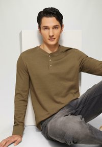 Camicia khaki a maniche lunghe con scollo henley e tre bottoni, abbinata a jeans grigi, seduta su uno sfondo bianco.