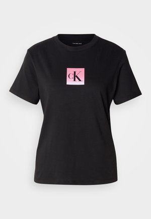 Črna bombažna majica s kratkimi rokavi z okroglim izrezom, ki prikazuje kvadratni rožnat gradient logotip z "cK" v črni pisavi na sredini.