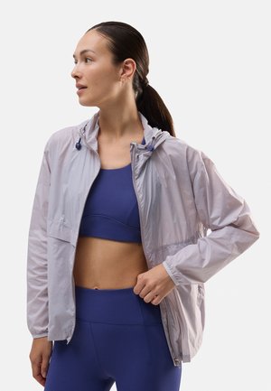 Chaqueta con capucha ligera y translúcida en un tono púrpura pálido, con cremallera frontal, puños elásticos y una silueta deportiva. Combinada con una blusa azul.