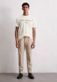 Cremfarbendes T-Shirt mit kurzen Ärmeln und "Marc O'Polo"-Druck, kombiniert mit beigen Hosen und schwarzen Slippern, vor einem hellen strukturierten Hintergrund.