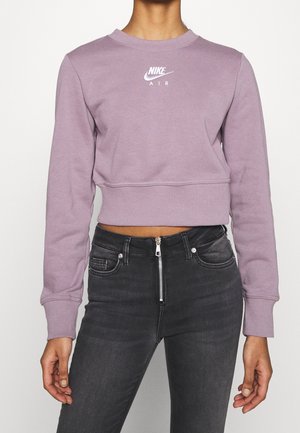 Osoba ubrana w krótką lawendową bluzę Nike Air oraz czarne jeansy z wysokim stanem z widocznym przednim zamkiem.