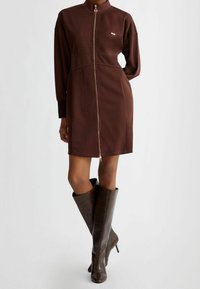 Brą dress z zamkiem błyskawicznym i wysokim kołnierzem, z długimi rękawami i fakturowanym materiałem; w połączeniu z ciemno brązowymi, wzorzystymi na skórę węża, botkami sięgającymi do kolan.