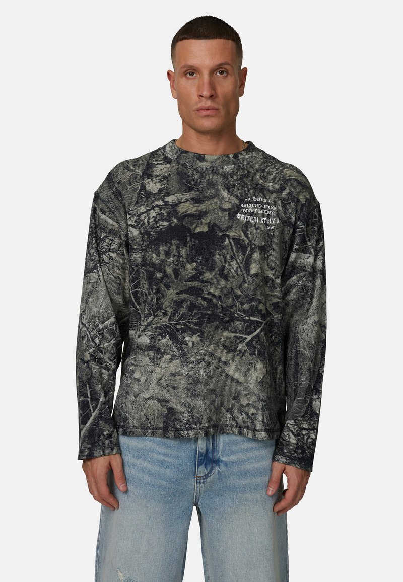 Homme portant une chemise à manches longues à motif camouflage sombre avec un texte blanc et un jean bleu clair, debout devant un fond uni.