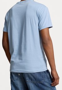 Person wearing a light blue short-sleeve T-shirt and blue denim jeans, shown from the back, standing against a plain background.