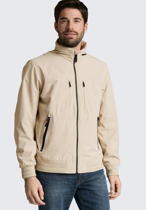 Uomo che indossa una giacca beige con cerniere sul petto e sui lati, abbinata a jeans blu, su sfondo chiaro uniforme.