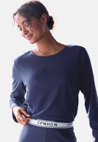 Tumesinise pika varrukaga särk, millel on rinnal süvistatud logo ja valge brändi vöö. Pehme, sile kangas, millel on kitsendatud siluett.