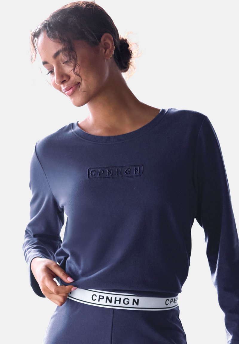 Tumesinise pika varrukaga särk, millel on rinnal süvistatud logo ja valge brändi vöö. Pehme, sile kangas, millel on kitsendatud siluett.