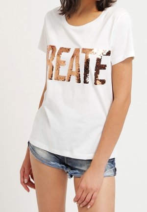 Frau trägt ein weißes T-Shirt mit Paillettenbuchstaben, die "REATE" buchstabieren, und Jeansshorts, steht mit einer Hand in der Tasche.