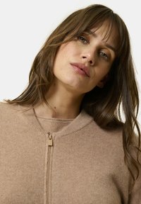Beige zip-up tröja med rund hals, mjuk textur och subtila sömdetaljer. Har en ljus tan dragkedja för stängning och en minimalistisk design.
