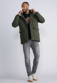Parka verde oliva con cierre de cremallera y a presión, dos bolsillos delanteros, capucha, llevada sobre un suéter negro y jeans grises, combinada con zapatillas blancas.