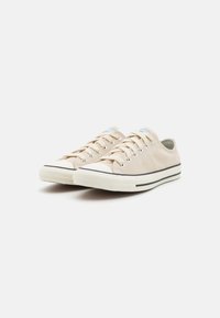 Beige canvas sneakers with white rubber toe cap, black stripe detail on sole, and laces. Smooth texture, low-top design.