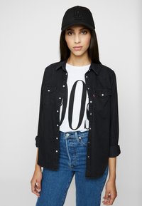 Cămașă din denim negru cu plăcuță de butoane, purtată peste un tricou alb cu litere negre îndrăznețe, asortată cu blugi bleu cu talie înaltă și o șapcă neagră.