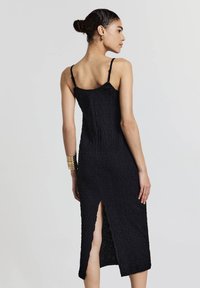 Schwarzes Midikleid aus strukturiertem Stoff, mit dünnen Trägern und einem Schlitz an der Seite; hat eine taillierte Silhouette, die knapp unter dem Knie endet.