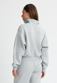 Sudadera gris con cuello alto y puños acanalados, corte relajado, textura suave y detalle de costura en la parte posterior, combinada con pantalones a juego.