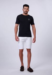 Must lühikeste varrukatega T-särk, millel on väike dekoratiivne logo, kombineeritud valgete lühikeste pükste ja tumedate pruunide libisevate kingadega. Lihtne ja vabaaja stiil.