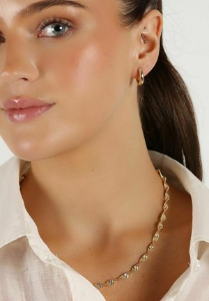 Gul halsband med en vriden design, tillsammans med små guldörhängen i form av hoops. Modellen har på sig en ljusfärgad skjorta med krage. Slät hudtextur.