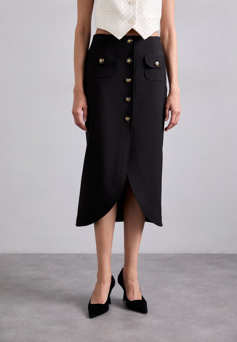 Jupe midi noire taille haute avec boutons dorés devant, poches à rabat, ourlet tulipe, portée avec un haut blanc sans manches et des talons pointus noirs.