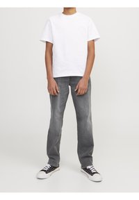 Persona de pie vistiendo una camiseta blanca sencilla, jeans gris rectos y zapatillas negras y blancas contra un fondo sencillo.