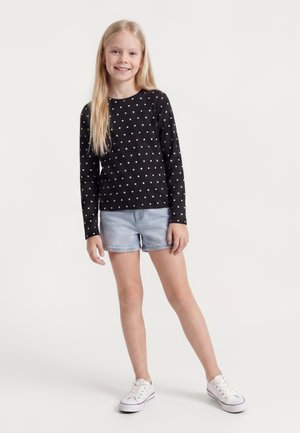 Blond pige smiler, iført sort langærmet prikket skjorte, lyse denimg shorts og hvide sneakers, stående på hvid baggrund.