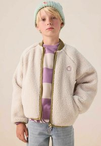 Blond kind draagt een lichtblauwe gebreide muts, een paarse en crèmekleurige gestreepte trui, een crèmekleurige fleece jas en lichtblauwe spijkerbroek, staand tegen een effen achtergrond.