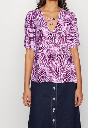 Vrouw draagt een paars-wit patroon blouse met strikdetail bij de halslijn en een donker denim rok met knoopsluiting aan de voorkant.