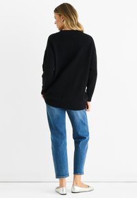 Černý oversized pletený svetr s kulatým výstřihem, kombinovaný s modrými džíny a plochými stříbrnými botami, zobrazený zezadu na bílém pozadí.