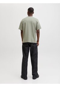 Άνδρας που στέκεται με την πλάτη προς το φακό φορώντας ανοιχτό πράσινο κοντομάνικο πουκάμισο, σκούρο τζιν και μαύρα παπούτσια σε απλό λευκό φόντο.