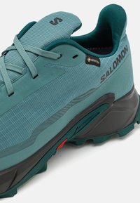 Turkusowe buty outdoorowe z teksturowaną górną częścią, wzmocnionym noskiem i czarną gumową podeszwą. Posiadają etykietę GORE-TEX oraz elementy odblaskowe.