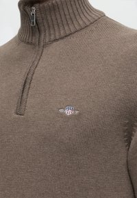 Pull marron en tricot avec col montant côtelé et fermeture éclair argentée, avec un petit logo brodé avec un bouclier et des étoiles sur la poitrine.