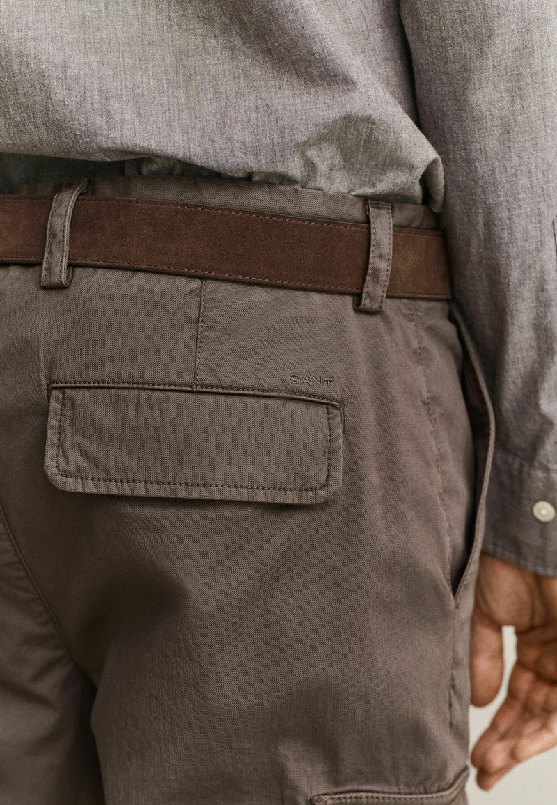 Vista trasera de una persona que lleva pantalones color topo con bolsillo con solapa y cinturón marrón, combinado con una camisa de manga larga gris claro.