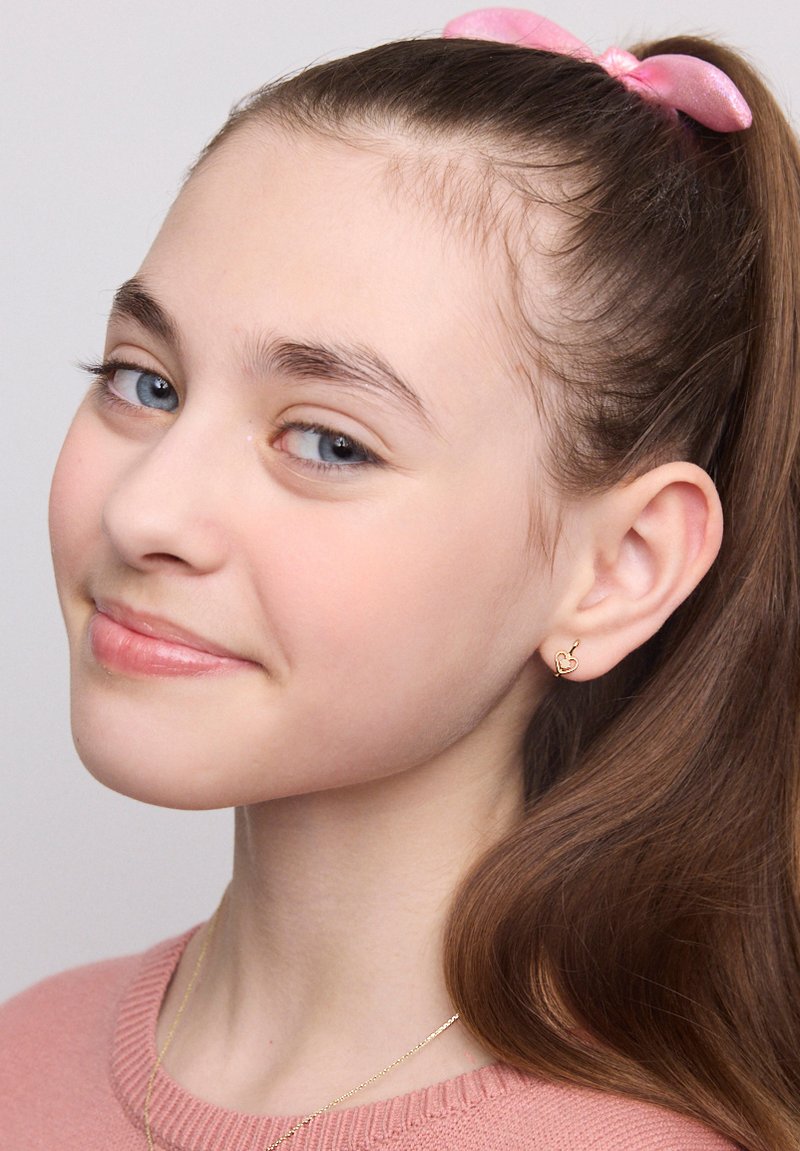 Złote kolczyki w kształcie serca, różowa wstążka we włosach, jasnobrązowe włosy, jasnoróżowy sweter, czyste niebieskie oczy, gładka skóra, delikatny rumieniec na policzkach.
